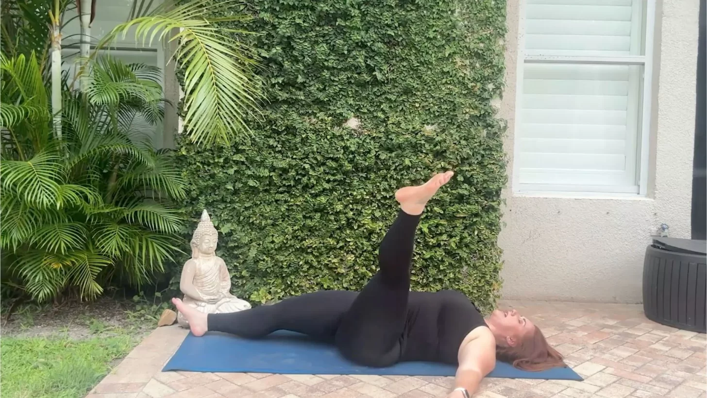 Женщина лежит на спине на коврике для йоги с одной ногой, вытянутой в воздухе.