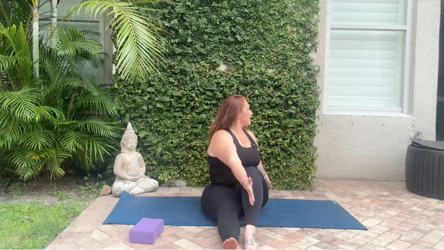 Женщина сидит на коврике для йоги с одной ногой прямой и одной ногой согнутой, скручиваясь в сторону.