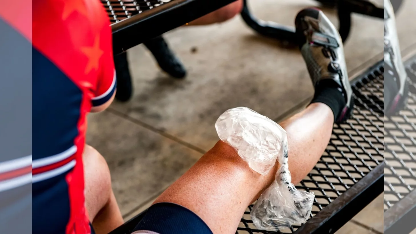 Мужчина-велосипедист в велоформе и обуви поднимает колено с пакетом льда.