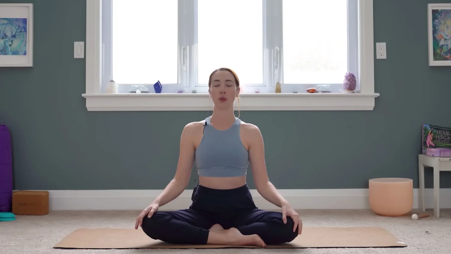 Yoga With Kassandra сидит в позе со скрещенными ногами, с руками на коленях и закрытыми глазами, практикуя йогу для снятия стресса.